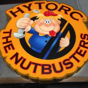May include: A circular illuminated sign with the text "HYTORC THE NUTBUSTERS" in red on a yellow background. It features a cartoon character holding a bolt and nut, wearing a red cap and blue shirt.