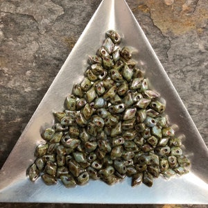 May include: A pile of light green and brown glass beads in a triangular tray. The beads are faceted and have a speckled appearance.