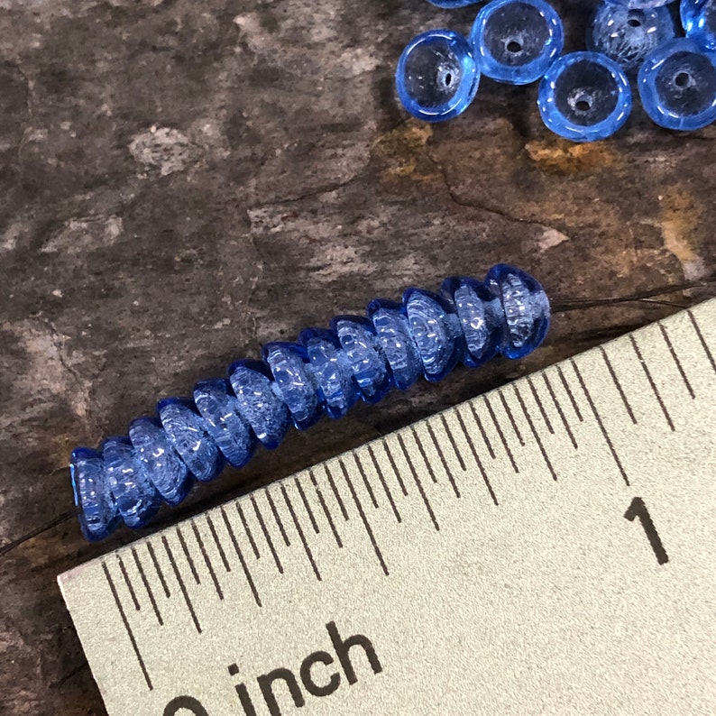 May include: A close-up of a strand of blue glass beads, measuring 1 inch in length. The beads are faceted and have a frosted finish. The beads are arranged in a spiral pattern.