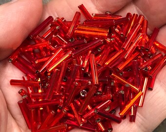 May include: A handful of vibrant, translucent red glass beads. These small, cylindrical beads are scattered, catching the light and reflecting a bright, fiery hue. Ideal for jewelry making or craft projects.
