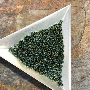 May include: A close-up of a pile of small, round, iridescent green beads in a triangular tray. The beads have a metallic sheen and are arranged in a random pattern.