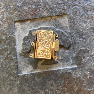 May include: Gold-toned metal jewelry clasp with a decorative floral design. The clasp has three loops for attaching jewelry.