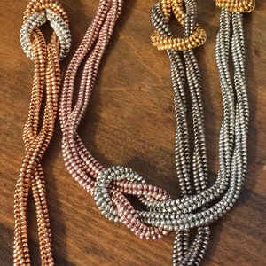 Könnte beinhalten: Eine mehrsträngige Halskette mit drei verschiedenen farbigen Metallketten. Die Ketten sind silber, gold und roségold. Die Halskette ist in einem dekorativen Muster verknotet.