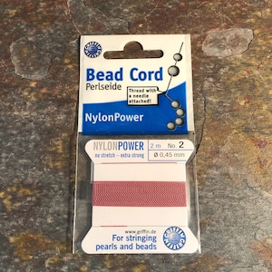 May include: A spool of red bead cord, labeled "Bead Cord Perlseide NylonPower" with a needle attached. The label also states "2 m No. 2 Ø 0,45 mm no stretch - extra strong".