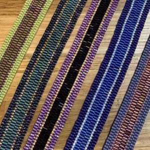 May include: Close-up of several beaded bracelets in various colors. The bracelets feature a variety of colors including green, navy, purple, black, blue, and red. The beads are arranged in rows, creating a textured surface. The bracelets are displayed on a wooden surface.