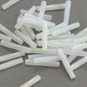 May include: A collection of white iridescent glass beads in a long, thin, cylindrical shape. The beads are scattered on a gray surface.