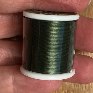 May include: A spool of dark green thread with a white label. The thread is wound tightly on the spool and has a slight sheen.