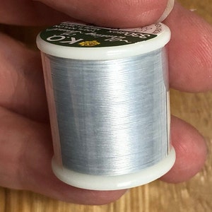 May include: A spool of light blue thread held between fingers. The spool has white plastic ends and a green and white label with text. The thread is labeled as K.O. Nylon.
