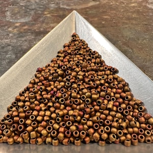 May include: A collection of small, cylindrical beads in shades of bronze and copper, arranged in a triangular silver tray. The beads have a metallic sheen and are densely packed, creating a textured surface. Ideal for jewelry making and crafting.