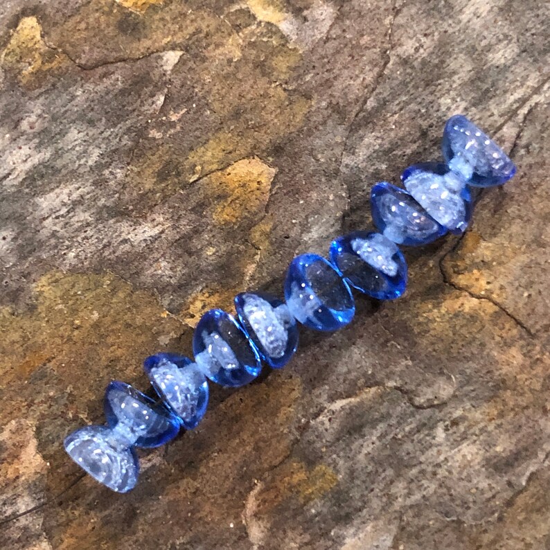 May include: A strand of light blue glass beads with a textured surface. The beads are shaped like teardrops and are strung together on a clear thread.