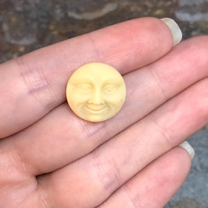 May include: A small, round, yellow button with a carved face. The face has closed eyes, a small nose, and a smiling mouth.