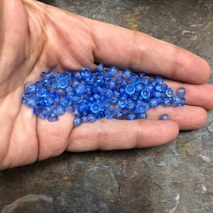 May include: A pile of small, light blue glass beads with a textured surface. The beads are round and have a slightly flattened top.