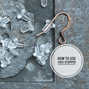 May include: A close-up of a pile of clear plastic earring stoppers. A copper earring hook is lying on top of the stoppers. A white circle with the text "HOW TO USE THIS STOPPER" is in the lower right corner of the image.