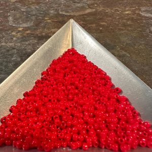 May include: A silver triangular tray filled with a large pile of small, bright red beads. The beads are densely packed, creating a vibrant, textured surface. The tray is set against a dark, textured background.