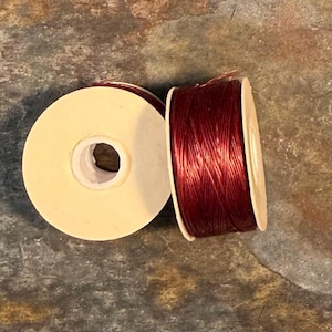 May include: Two spools of burgundy-colored thread. One spool is full of thread, while the other is empty. The spools have a cream-colored cardboard core. The thread appears to be tightly wound and ready for use.