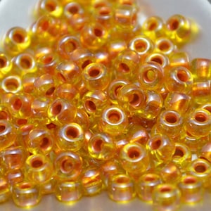 May include: A pile of small, transparent, yellow and orange glass beads. Each bead has a hole running through the center. The beads are scattered, creating a textured surface. The beads have a shiny, reflective quality.