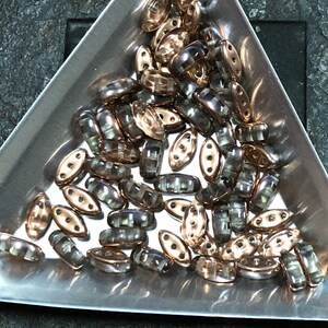 May include: A collection of oval-shaped glass beads with a metallic finish. The beads have a translucent, iridescent appearance with a mix of gold and gray tones.