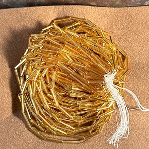 May include: A large quantity of gold-colored glass beads in a tube shape. The beads are bundled together and tied with a white string.