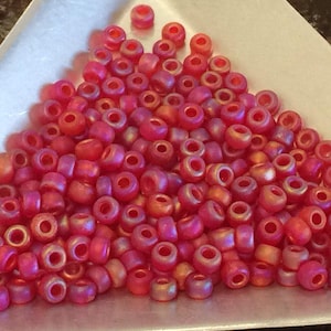 May include: A pile of small, red beads with a metallic sheen. The beads are cylindrical with a hole running through the center. The color shifts from red to orange in the light. These beads are suitable for jewelry making and crafts.