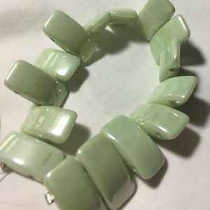 May include: A light green bracelet composed of rectangular beads. The beads are strung together to form a flexible band. The bracelet is a pale, muted green color and has a glossy finish. The beads are approximately 1 inch long.