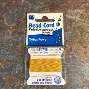 May include: A spool of gold nylon bead cord, labeled "NylonPower Bead Cord Perlseide" with the text "Thread with a needle attached!" and "For stringing pearls and beads". The spool is 2 meters long and 0.80 millimeters in diameter.