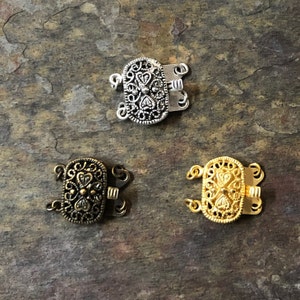 May include: Three ornate metal clasps for jewelry making. The clasps are silver, bronze, and gold in color. Each clasp has a decorative design with a heart shape in the center.