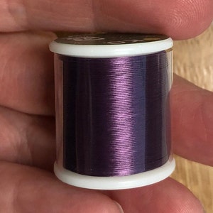 May include: A spool of metallic purple thread in a clear plastic container with a white lid.