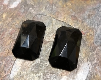May include: Two black faceted glass cabochons, each with a rectangular shape and a slightly rounded edge. The cabochons are set against a rough, gray stone background.