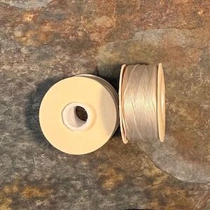 May include: Two spools of thread are displayed. One spool is a light beige color, and the other is a light beige thread wound around a wooden spool. The spools are on a textured, multi-colored surface.