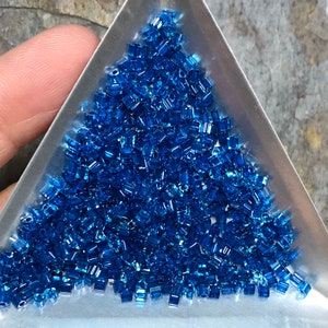 May include: A close-up view of a pile of small, blue, glass beads. The beads are in a triangular-shaped container.