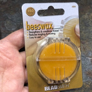 May include: A package of beeswax for strengthening and conditioning thread, reducing tangling and knotting. The package has a clear plastic container holding a round, yellow wax disc. The text "beeswax" is prominently displayed.