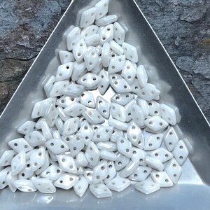 May include: A close-up of a pile of white, iridescent, two-hole, diamond-shaped beads.