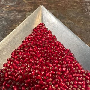 May include: A silver metal tray filled with a mound of small, red seed beads. The beads have a translucent quality, revealing a golden interior. The tray is positioned on a textured, dark surface.