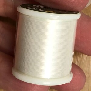 May include: A white spool of thread with a clear plastic container. The container has a black label with the text "24/0" printed on it.