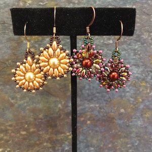 May include: Two pairs of beaded earrings displayed on a black stand. One pair features a golden sunflower design, while the other showcases a cluster of red, green, and pink beads. The earrings have gold and copper-colored ear wires.