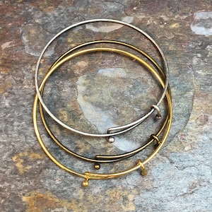 May include: Three bangle bracelets, one silver, one gold, and one brass, stacked on top of each other. The bracelets are all open and have a simple design.