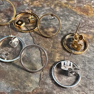May include: A set of four gold and silver hoop earrings with post backs. The earrings are all different sizes and are arranged on a textured brown surface.