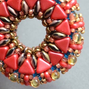 May include: A close-up of a red, gold, and blue beaded jewelry piece. The piece is made of small, triangular beads and round beads. The beads are arranged in a circular pattern.