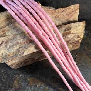 May include: A strand of pink glass beads, each bead is a cylinder made of stacked discs. The beads are arranged in a row on a piece of wood.
