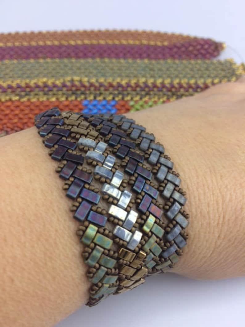 May include: A multi-colored beaded bracelet with a geometric pattern. The bracelet is made with small, square beads in shades of brown, green, blue, and silver.