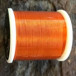 May include: A spool of orange thread with white plastic sides. The thread is tightly wound around the spool, and the color is a vibrant, warm orange. The spool has a black label with white text.