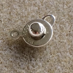 May include: Silver-toned metal clasp with a round button and a loop. The button has a raised center and the words "Prym" are engraved on the side.