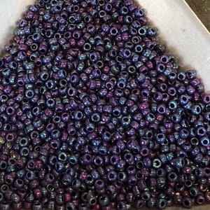 May include: A pile of small, dark purple beads with a metallic sheen. The beads are round with a hole through the center, and they appear to be made of glass or a similar material. The beads are scattered across a light-colored surface.