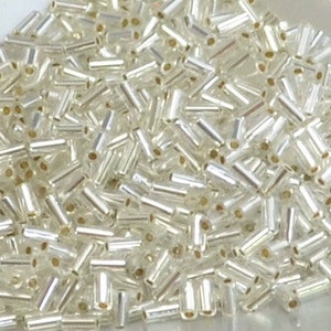 May include: A close-up shot of a pile of small, clear, cylindrical beads with a silver-like coating. The beads are scattered, showing their tubular shape and the holes running through them. The beads are likely used for jewelry making or crafts.