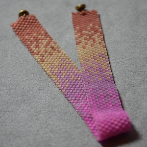 May include: A pink, beige, and peach beaded bracelet with gold metal clasps. The bracelet is made with small, square beads that are woven together in a pattern.