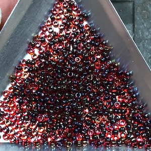 May include: A close-up shot of a collection of small, round beads in a silver tray. The beads are primarily a deep red color with hints of iridescent purple and gold. The beads are densely packed together.