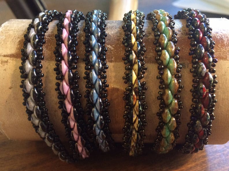 Op de afbeelding: Zes kralenarmbanden in verschillende kleuren en patronen. De armbanden zijn gemaakt van zwarte kralen en andere gekleurde kralen. De armbanden zijn in een rij op een bruin oppervlak gerangschikt.