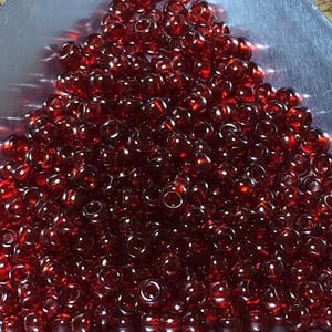 May include: A pile of small, transparent, dark red glass beads. The beads are round with a hole through the center, and they are scattered across a silver surface. The beads have a glossy finish and reflect light.