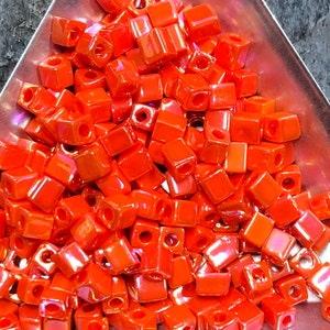 May include: A large pile of small, orange, square beads with a iridescent finish. The beads are arranged in a random pattern.