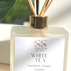 May include: A clear glass bottle with a gold lid and wooden sticks, filled with a white tea scented reed diffuser. The label reads "White Tea" with the scent notes "Mandarin Ginger Jasmine" and "SCENTS FOR THE SOUL". It is hand poured in Charlotte, NC and is 6 oz (177 ml).